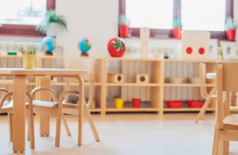 Leerer Kindergartenraum mit Holztischen, Stühlen und Regalen voller Spielzeug, Globen und Pflanzen vor großen Fenstern.