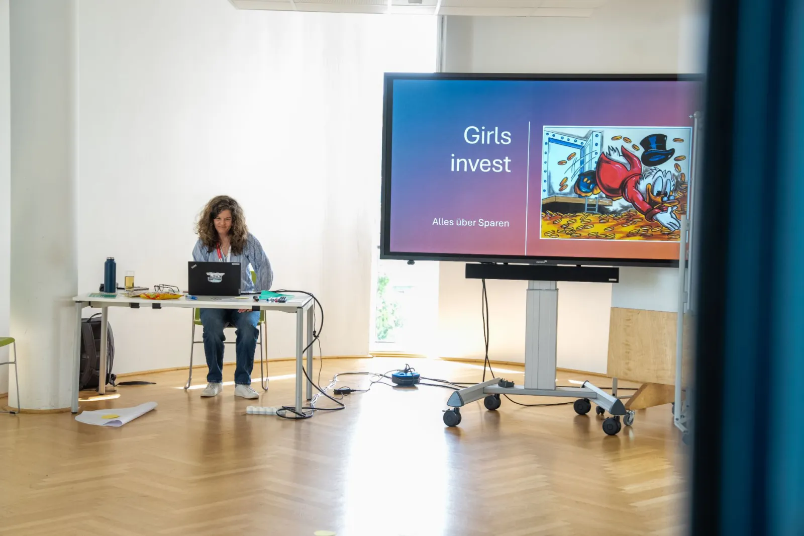 Person sitzt an Schreibtisch mit Laptop; großer Bildschirm zeigt Folie 'Girls invest' und 'Alles über Sparen' in Seminarraum.