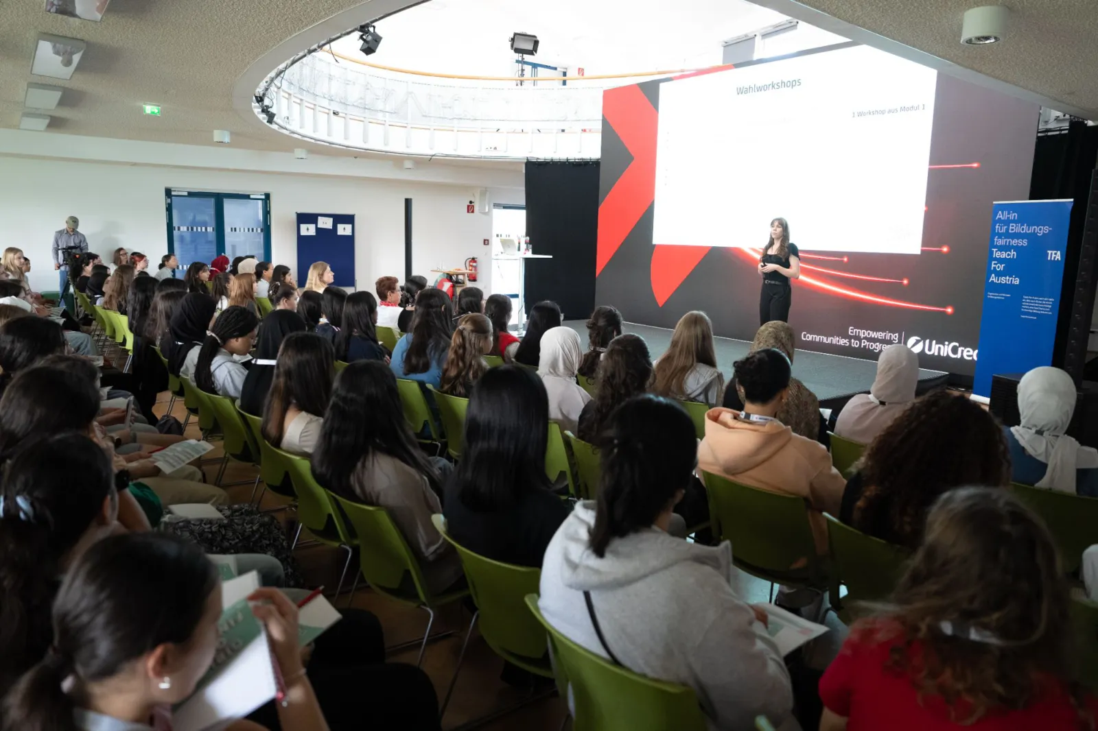 Person hält Vortrag vor Publikum in Konferenzsaal; große Bildschirmprojektion mit Folien im Hintergrund.