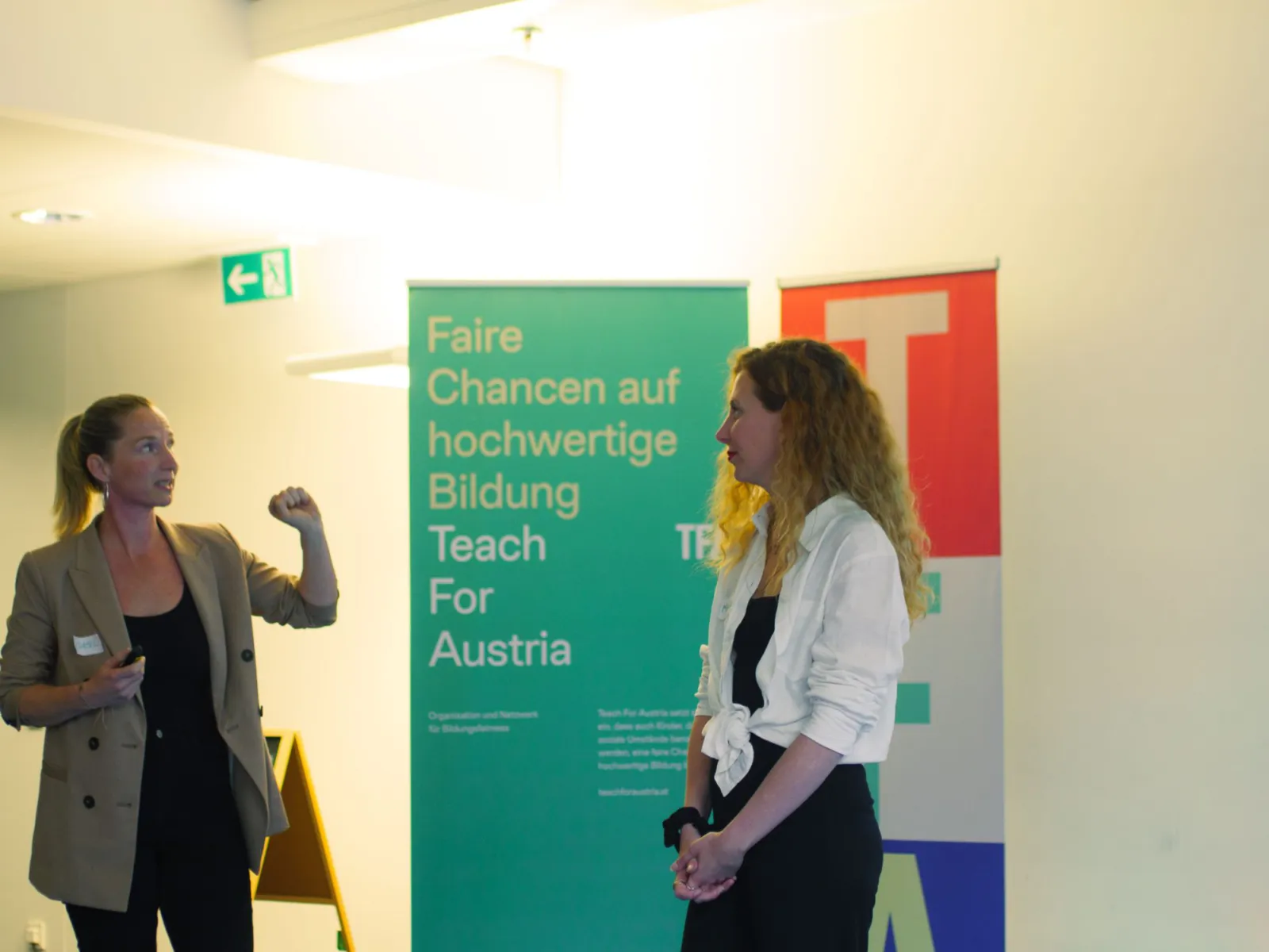 Zwei Personen im Gespräch auf Bildungsmesse; linke Person hebt Faust, rechte lauscht; Banner zeigt Faire Chancen auf hochwertige Bildung.