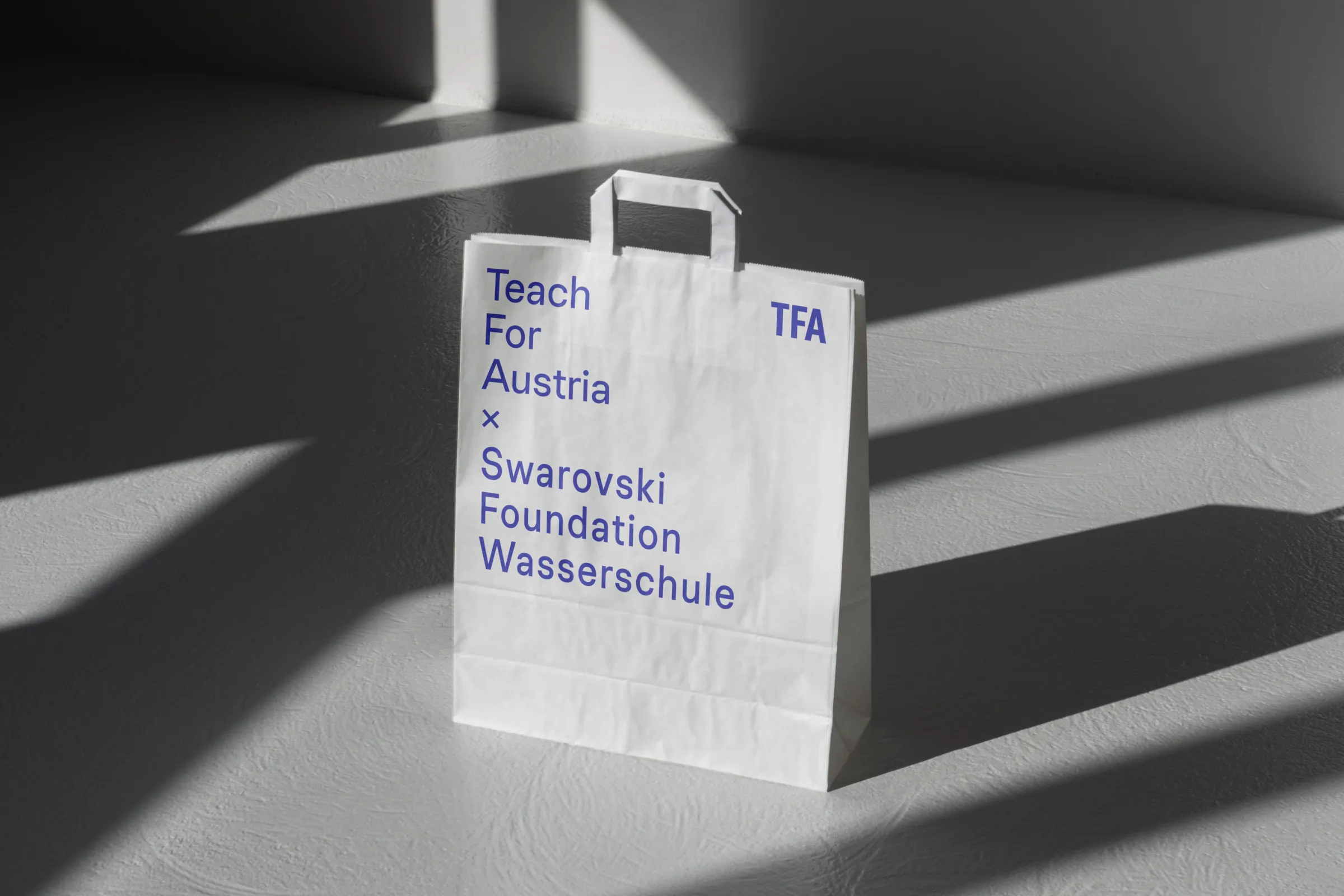 Weiße Tragetasche mit blauem Text 'Teach For Austria x Swarovski Foundation Wasserschule' steht auf grauem Boden; Schatten von Fensterjalousien.