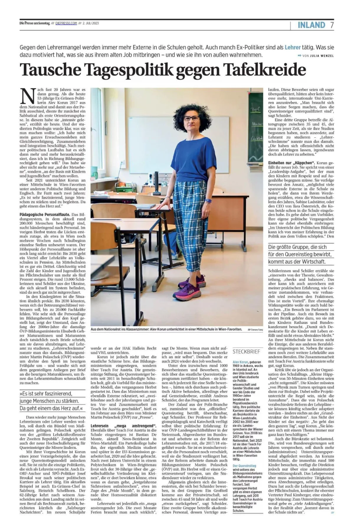 Lehrkraft steht im Klassenzimmer vor Schreibtischen, trägt blaues Oberteil und karierte Hose.