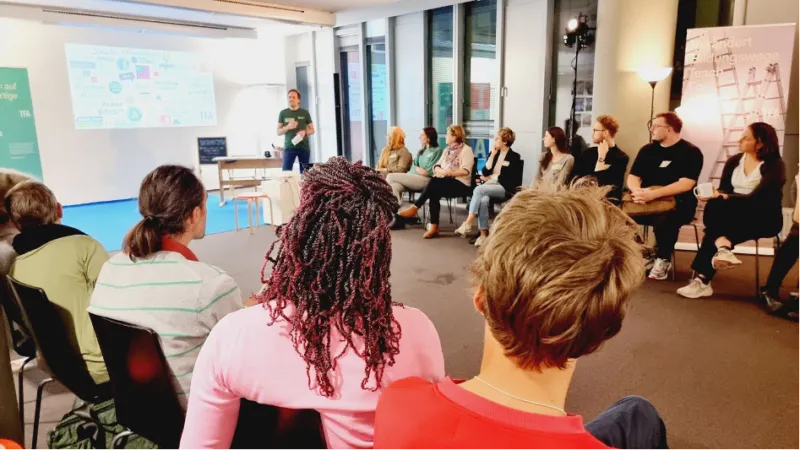 Person steht vor Projektionswand und hält Vortrag, während Teilnehmende im Seminarraum zuhören.