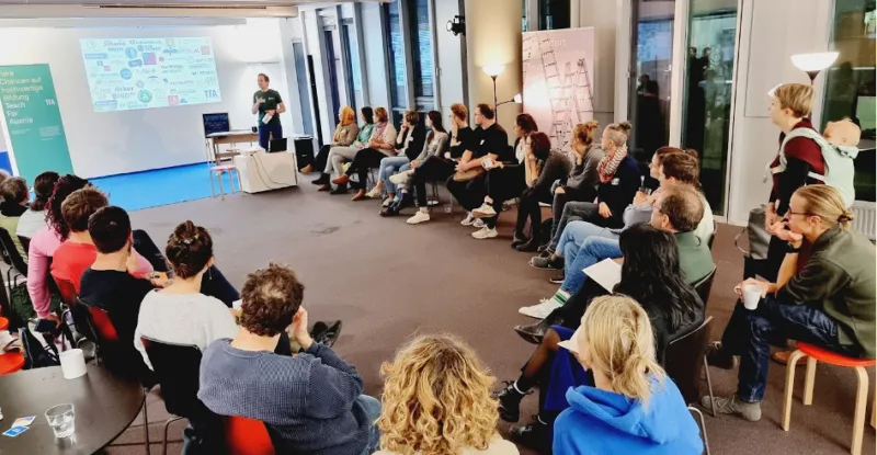 Referent hält Vortrag vor Präsentationsleinwand mit Diagrammen; Publikum sitzt in Halbkreis in modernem Konferenzraum.