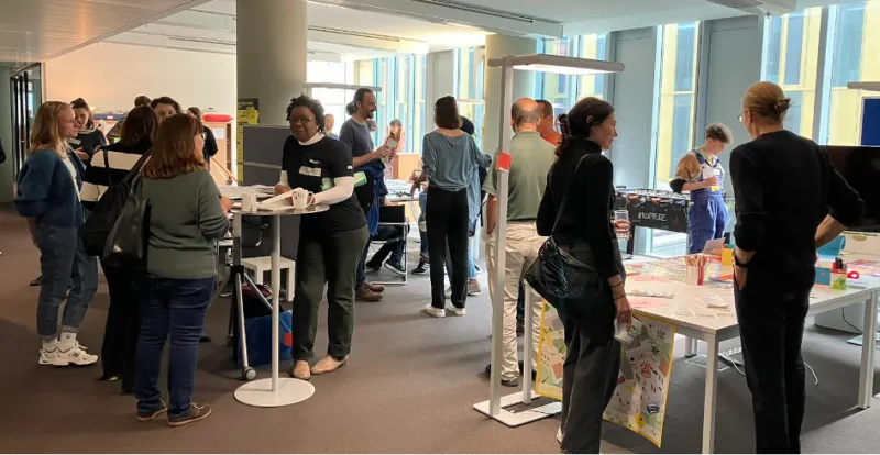 Teilnehmende stehen an mehreren Stehtischen und erkunden interaktive Stände in einem hellen Konferenzraum.