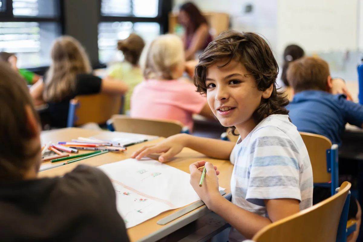 Kind zeichnet mit grünem Buntstift an Tisch im Klassenraum; weitere Kinder sitzen im Hintergrund.