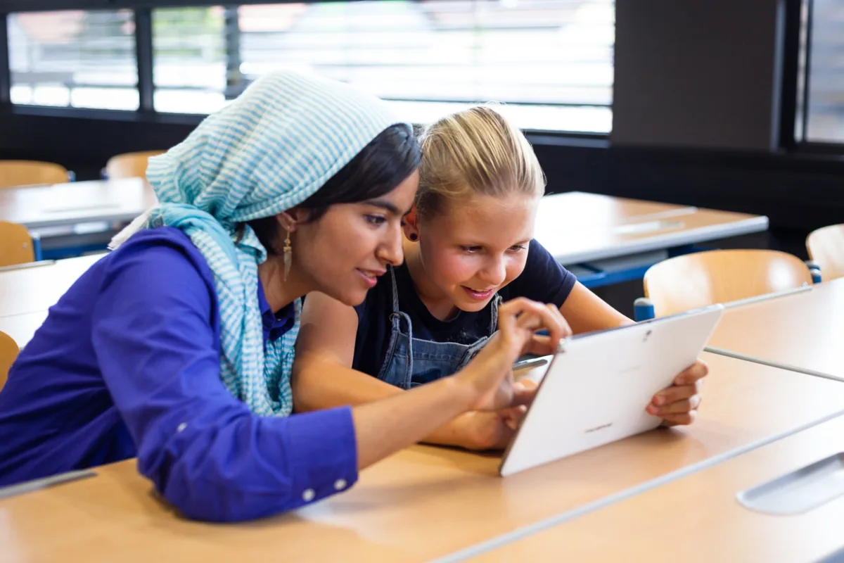 Zwei Personen arbeiten gemeinsam an einem Tablet in einem Klassenraum.