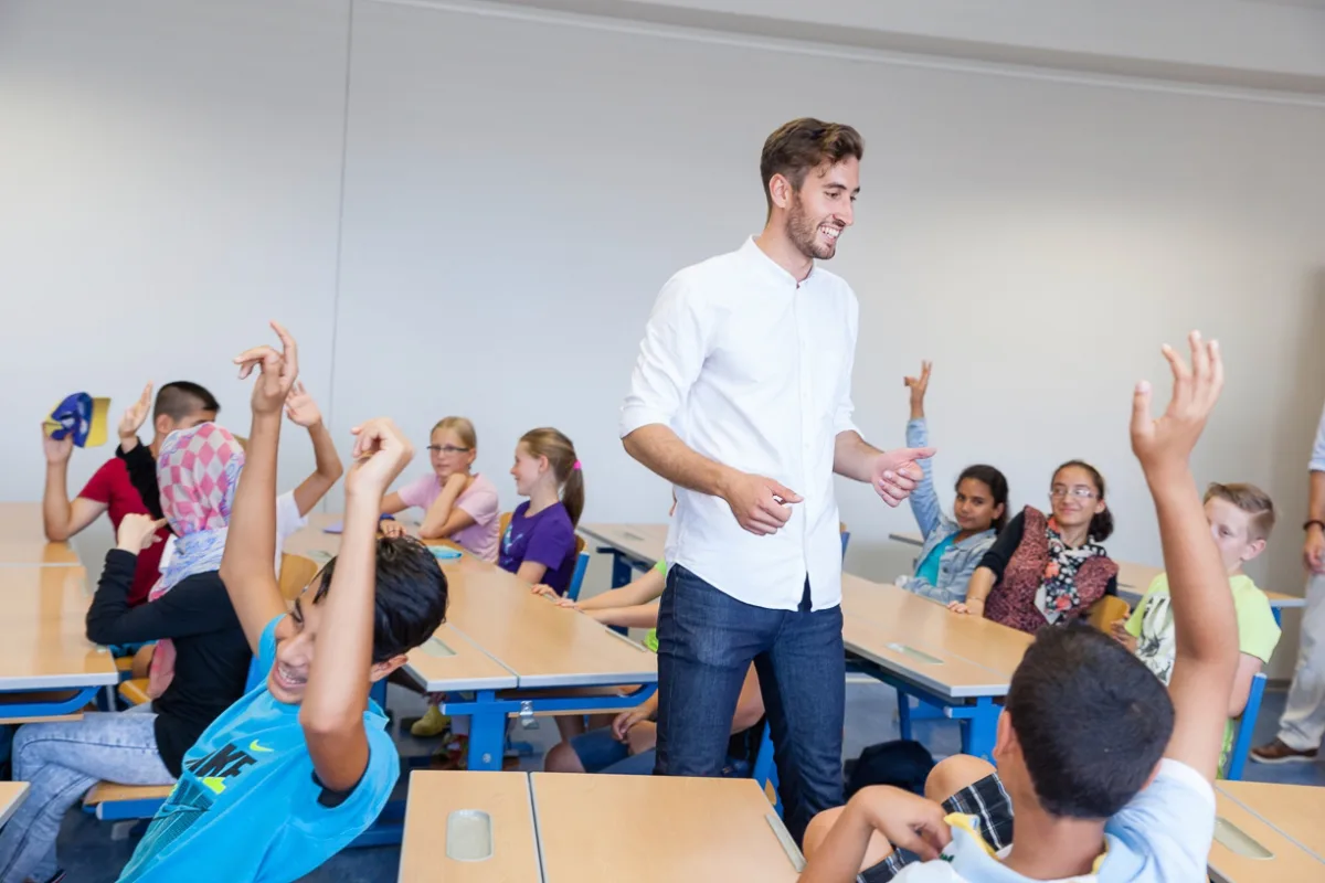 Lehrperson steht im Klassenzimmer und unterrichtet Gruppe von Lernenden; viele Hände sind erhoben.