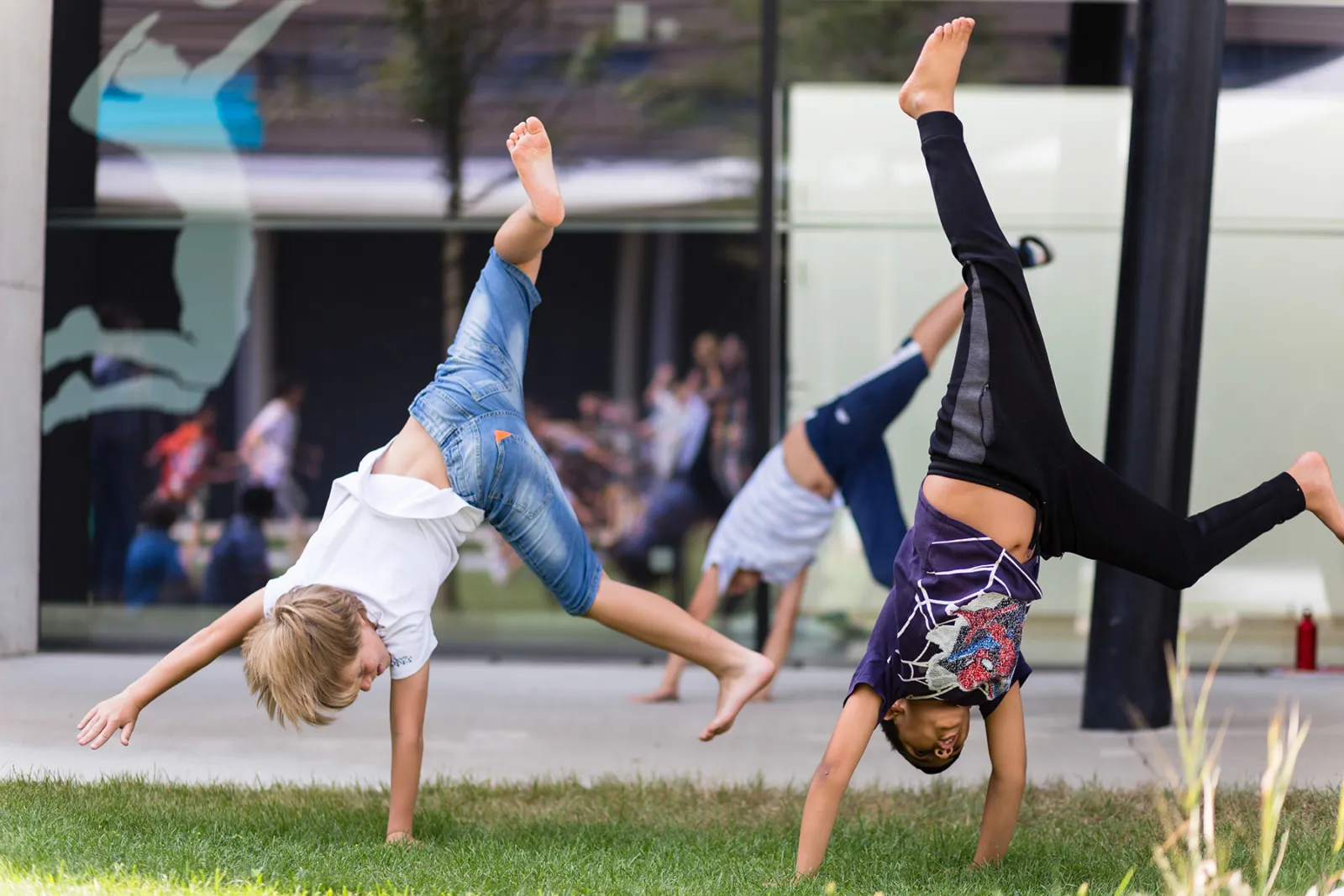Zwei Kinder machen Handstände auf der Rasenfläche vor moderner Glasfassade; weitere Kinder im Hintergrund.