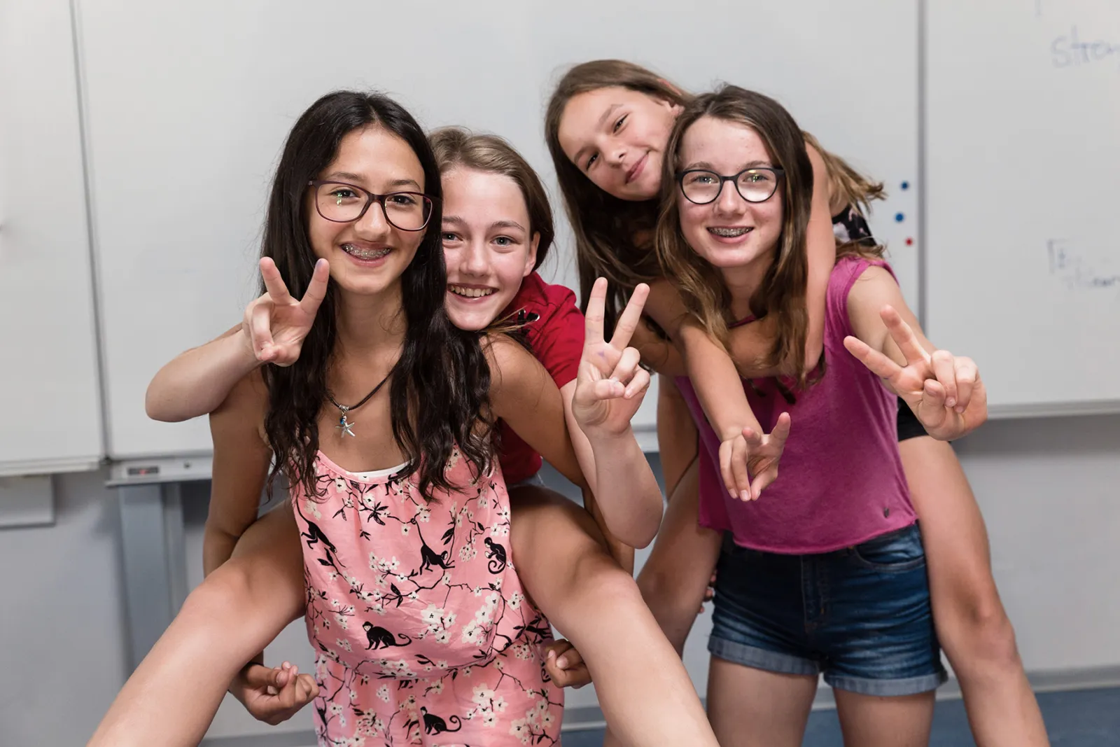 Vier Mädchen machen ein Selfie, zeigen Peace-Zeichen, im Klassenzimmer vor Whiteboard.