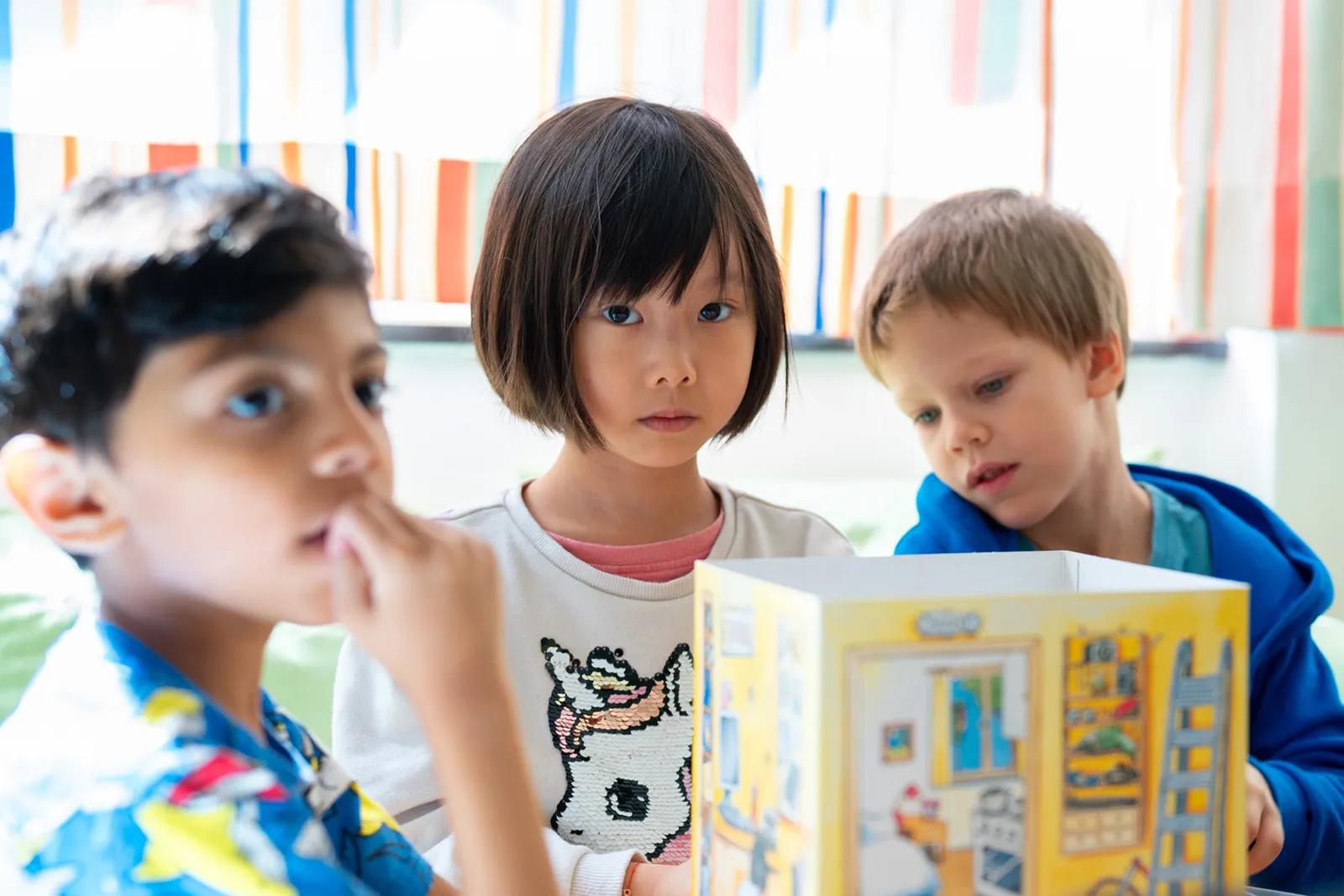 Drei Kinder betrachten ein gelbes Spielzeughaus; zentrale Person in der Mitte, flankiert von zwei Jungen.