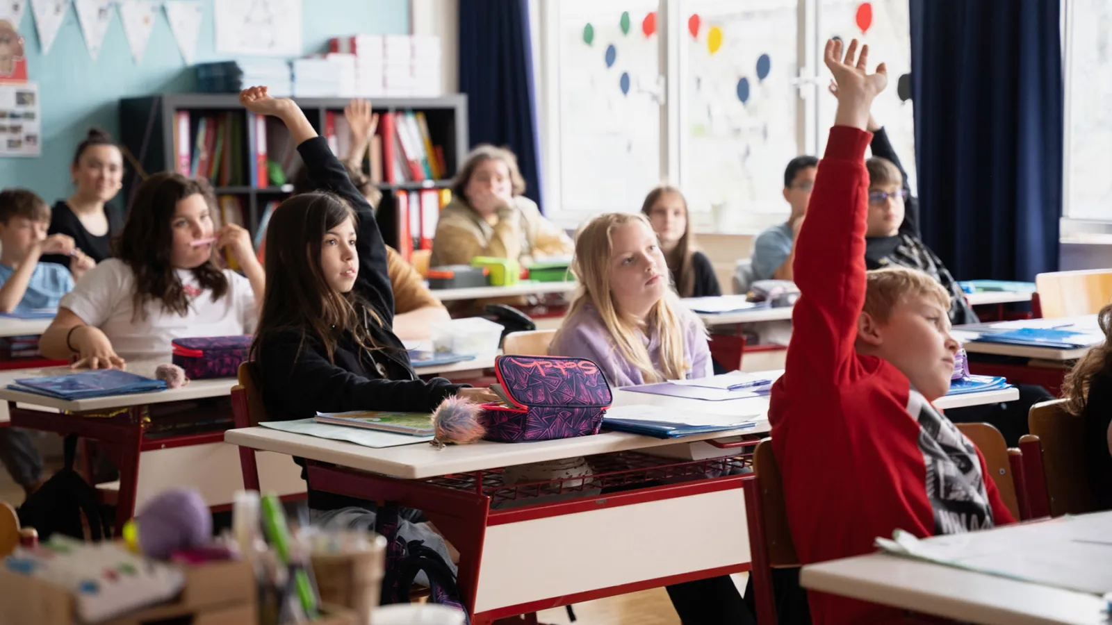 Schülerinnen und Schüler im Klassenraum heben die Hände; Lernmaterialien liegen auf Tischen.