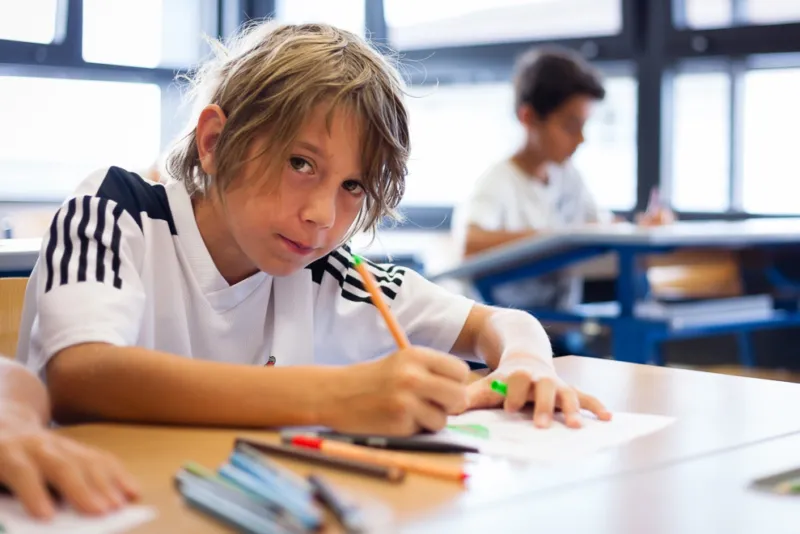 Schüler:in zeichnet mit Bleistift auf Arbeitsblatt am Schultisch; Klassenraum im Hintergrund.