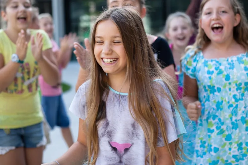 Mädchen mit langen braunen Haaren lächelt, umgeben von applaudierenden Kindern draußen.