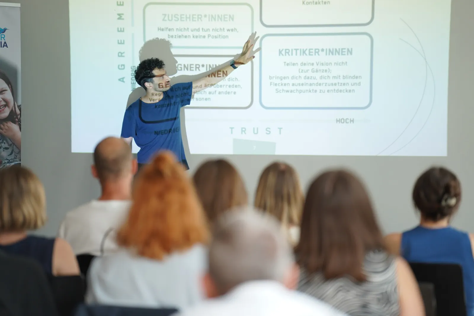 Person in blauem T-Shirt erklärt Diagramm an der Leinwand vor Publikum im Seminarraum.
