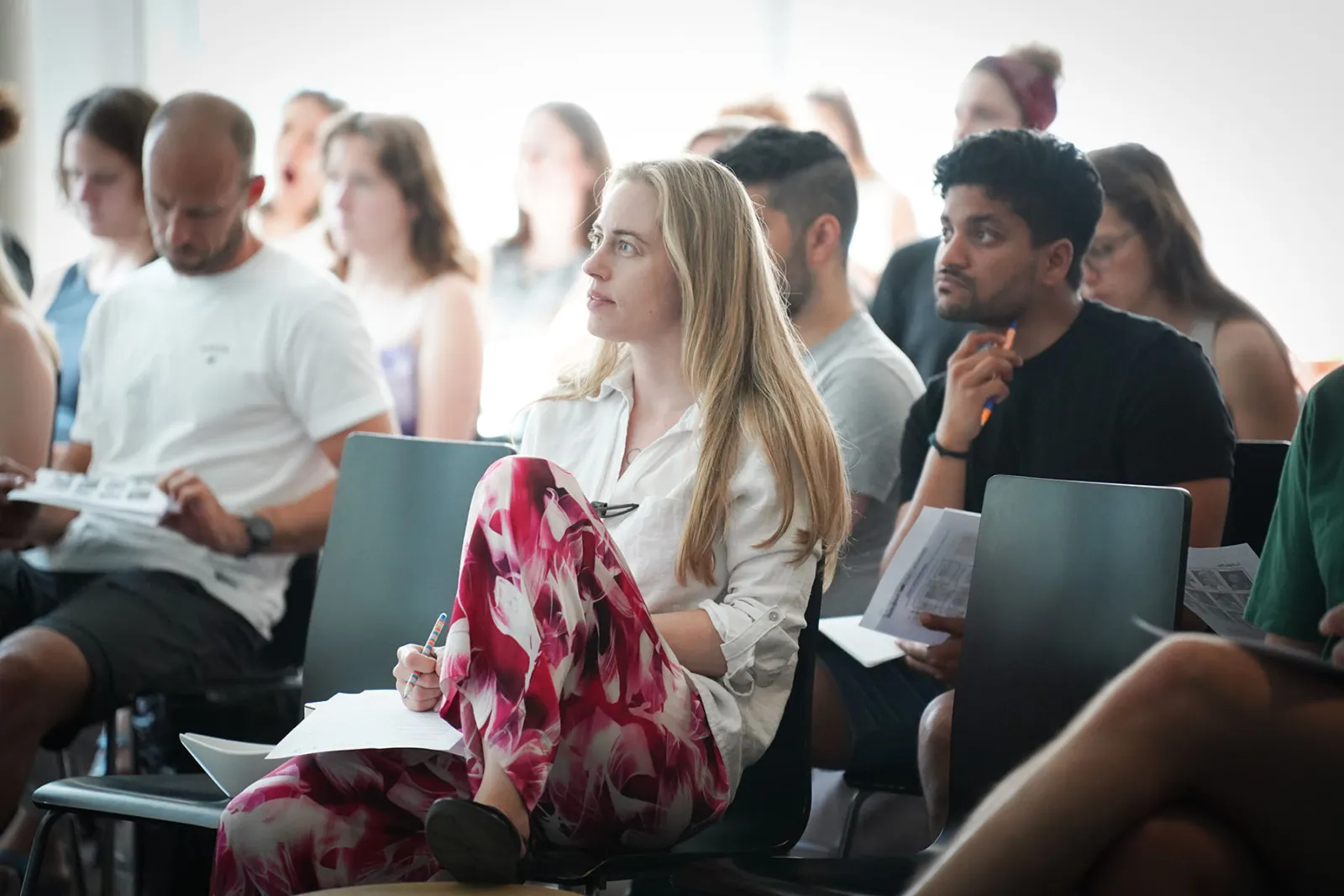 Person mit blonden Haaren in weißem Hemd sitzt in Seminar, notiert auf Papier, pink-weiß gemusterte Blumenhose; Hintergrund mit weiteren Teilnehmenden.