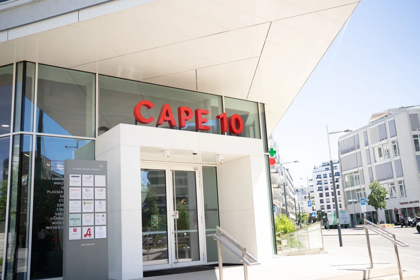 Moderne Gebäudefassade mit Glasfront und rotem CAPE 10 über dem Eingang; Straßenzug im Hintergrund.