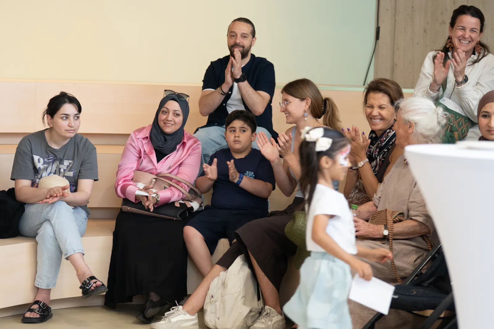 Vielfältige Gruppe von Erwachsenen und Kindern sitzt auf Holzbänken in einem Innenraum, klatscht; ein Kind mit Zettel geht vorbei.