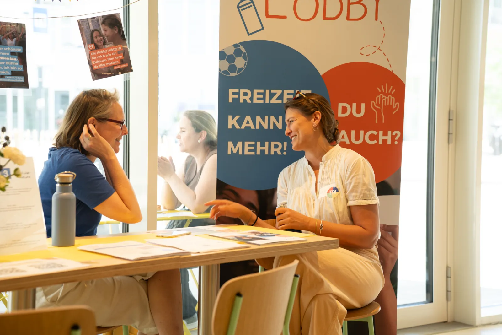 Zwei Personen unterhalten sich an einem Beratungstisch in einem hellen Raum; großes Banner im Hintergrund.
