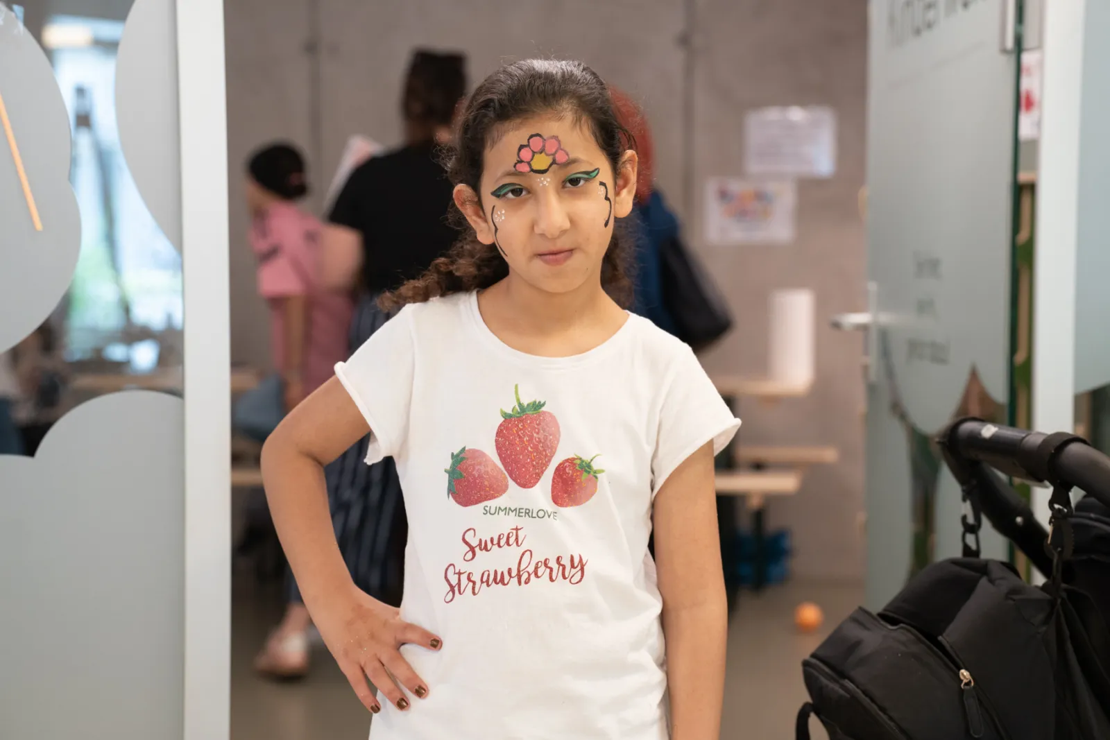Mädchen mit Gesichtsbemalung (Stirnblume) trägt weißes T-Shirt mit Erdbeer-Motiv und steht in Innenraum, eine Hand auf der Hüfte.
