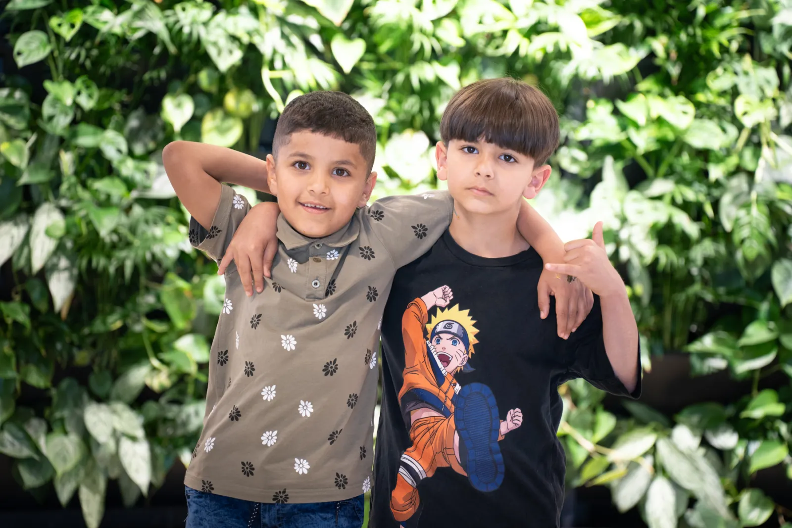 Zwei Kinder stehen eng, Arme um die Schultern; eines trägt Naruto-T-Shirt, das andere Polohemd mit Blumenmuster; grüne Pflanzenwand.