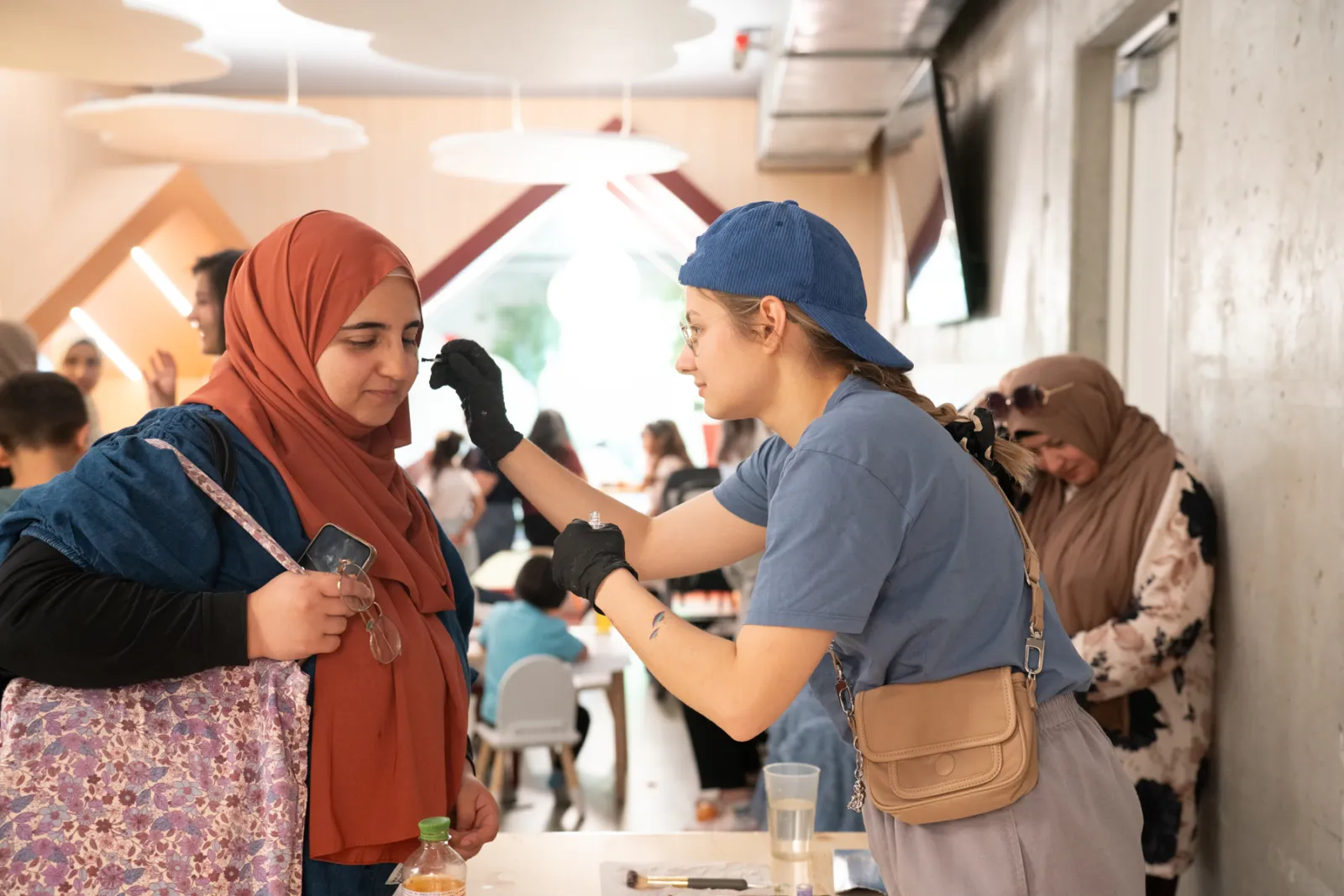 Visagistin in blauem T-Shirt und blauer Kappe schminkt eine Frau mit orangefarbenem Hijab in einem Veranstaltungsraum.