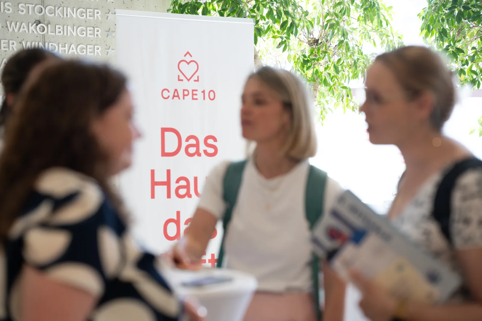 Drei Personen an einem Informationsstand; eine Person reicht einer anderen die Hand, vor Cape10 Banner mit Logo und grünem Laub im Hintergrund.