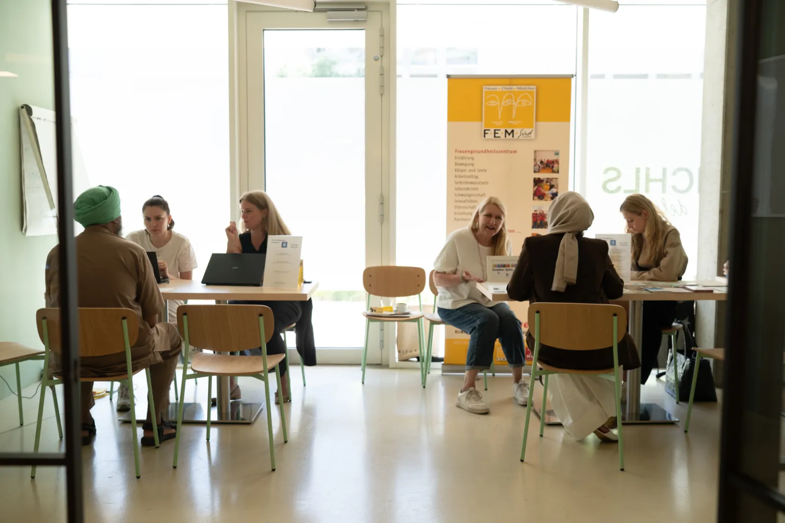 Mehrere Personen führen Beratungsgespräche an Tischen in hellem Innenraum; Hintergrundbanner sichtbar.