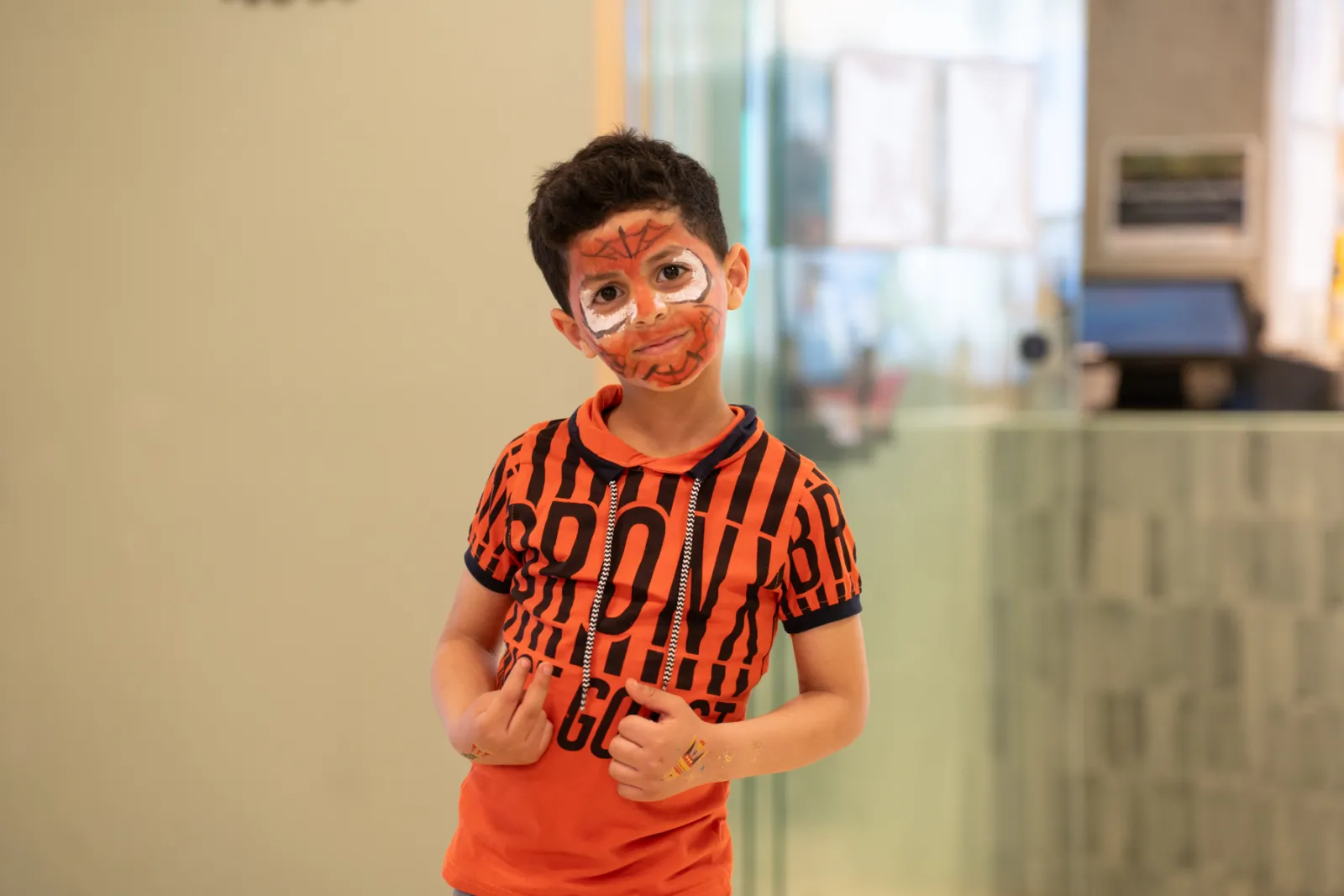 Kind mit Tiger-Gesichtsbemalung, orangefarbenem T-Shirt mit schwarzem Muster, steht in Innenraum und schaut zur Kamera.