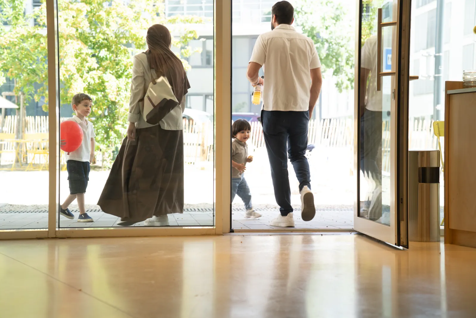 Familie geht durch offene Glastüren eines hellen Innenraums; draußen spielen Kinder mit Ball.