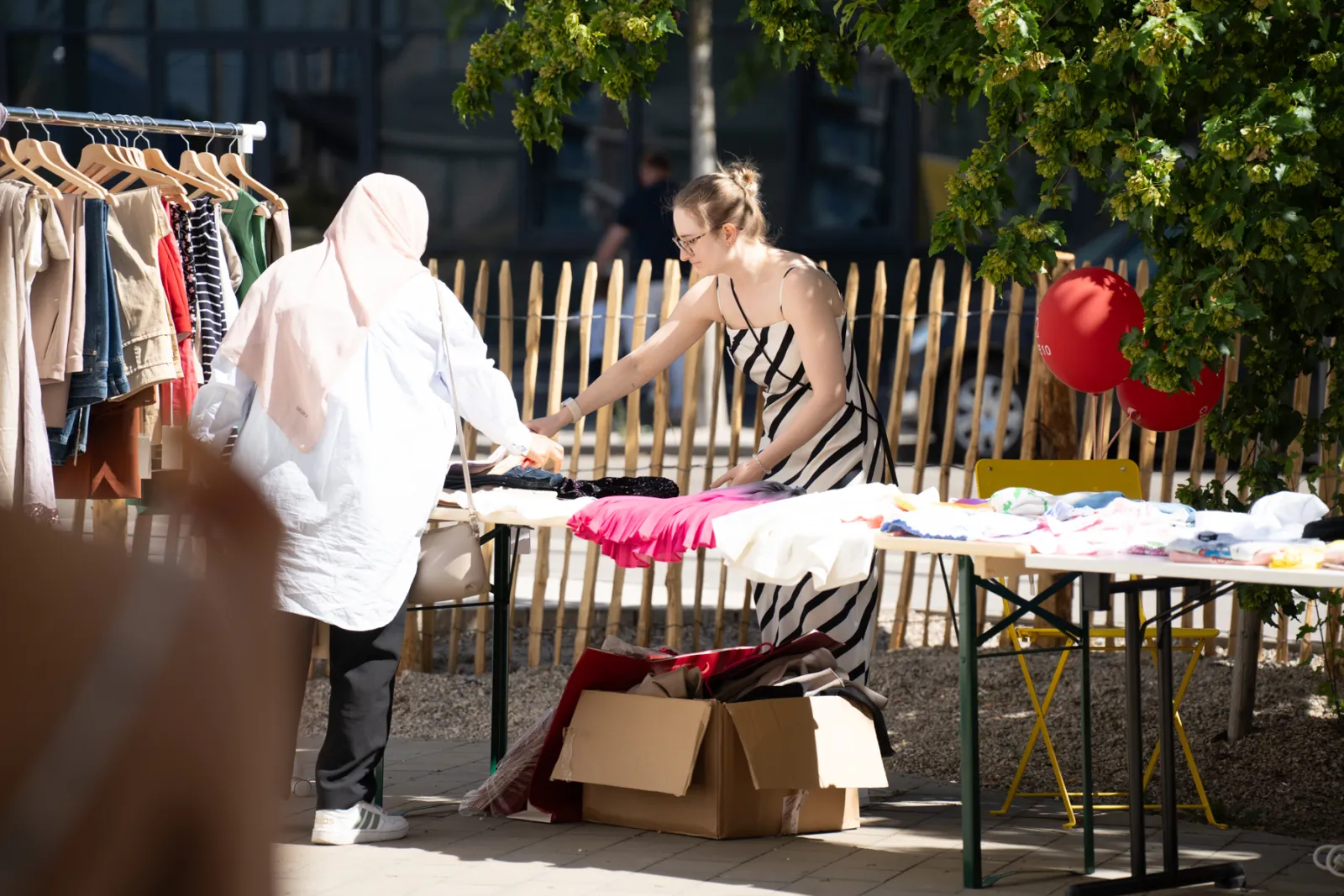 Zwei Frauen an einem offenen Kleidungsstand; eine Person mit Hijab reicht Kleidung über einen Tisch; Kartons darunter, Zäune und Bäume im Hintergrund.