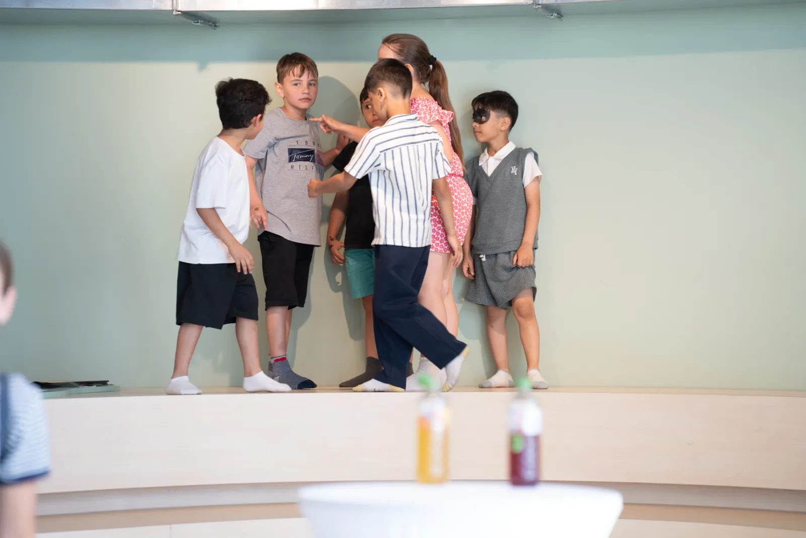 Gruppe von Kindern auf einer Bühne; mehrere stehen im Gespräch, andere beobachten; grüne Wand im Hintergrund; Getränke unscharf im Vordergrund.