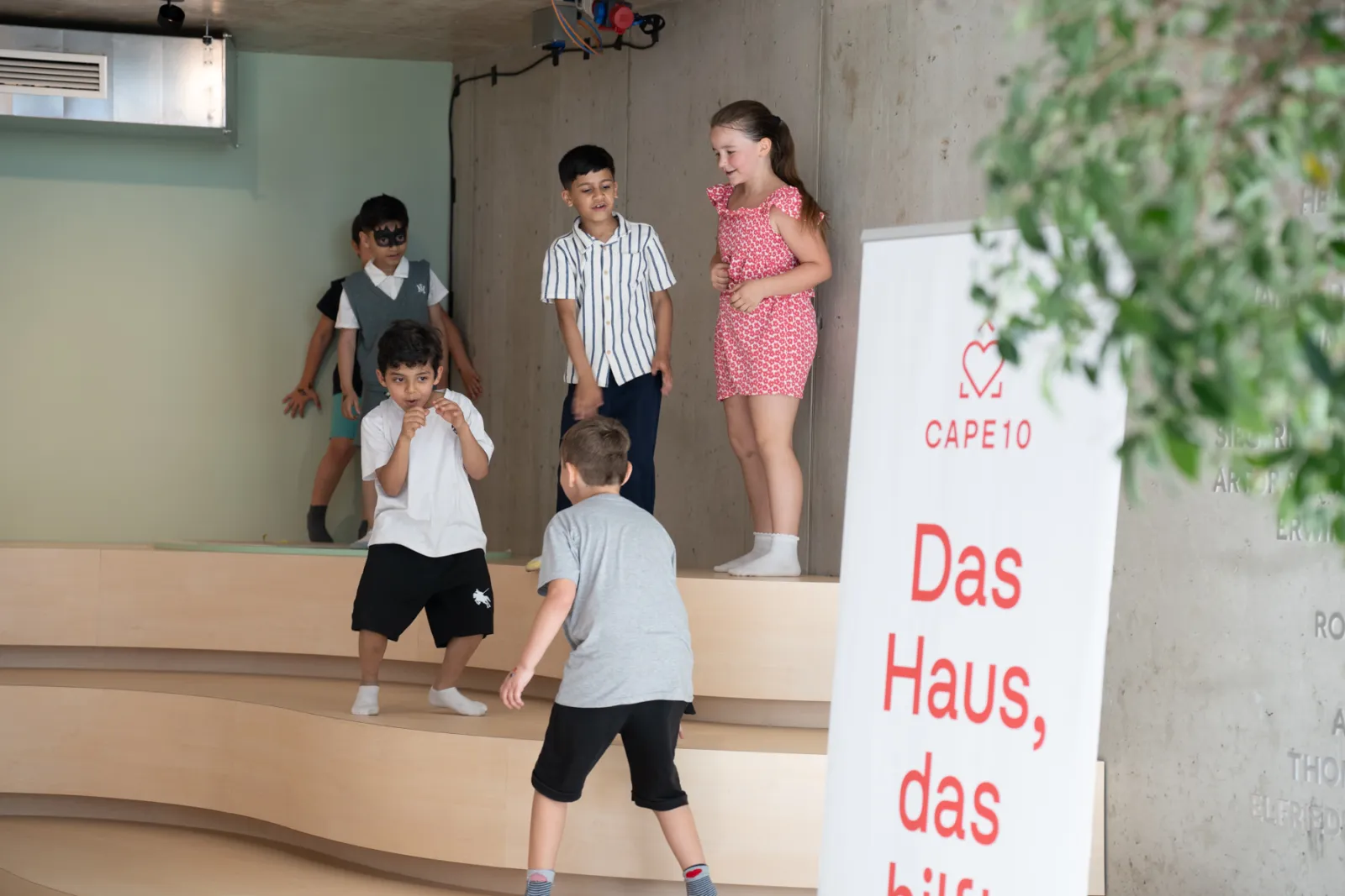 Gruppe von Kindern auf einer mehrstufigen Holztreppe in einem Innenraum, einige stehen, andere klettern.