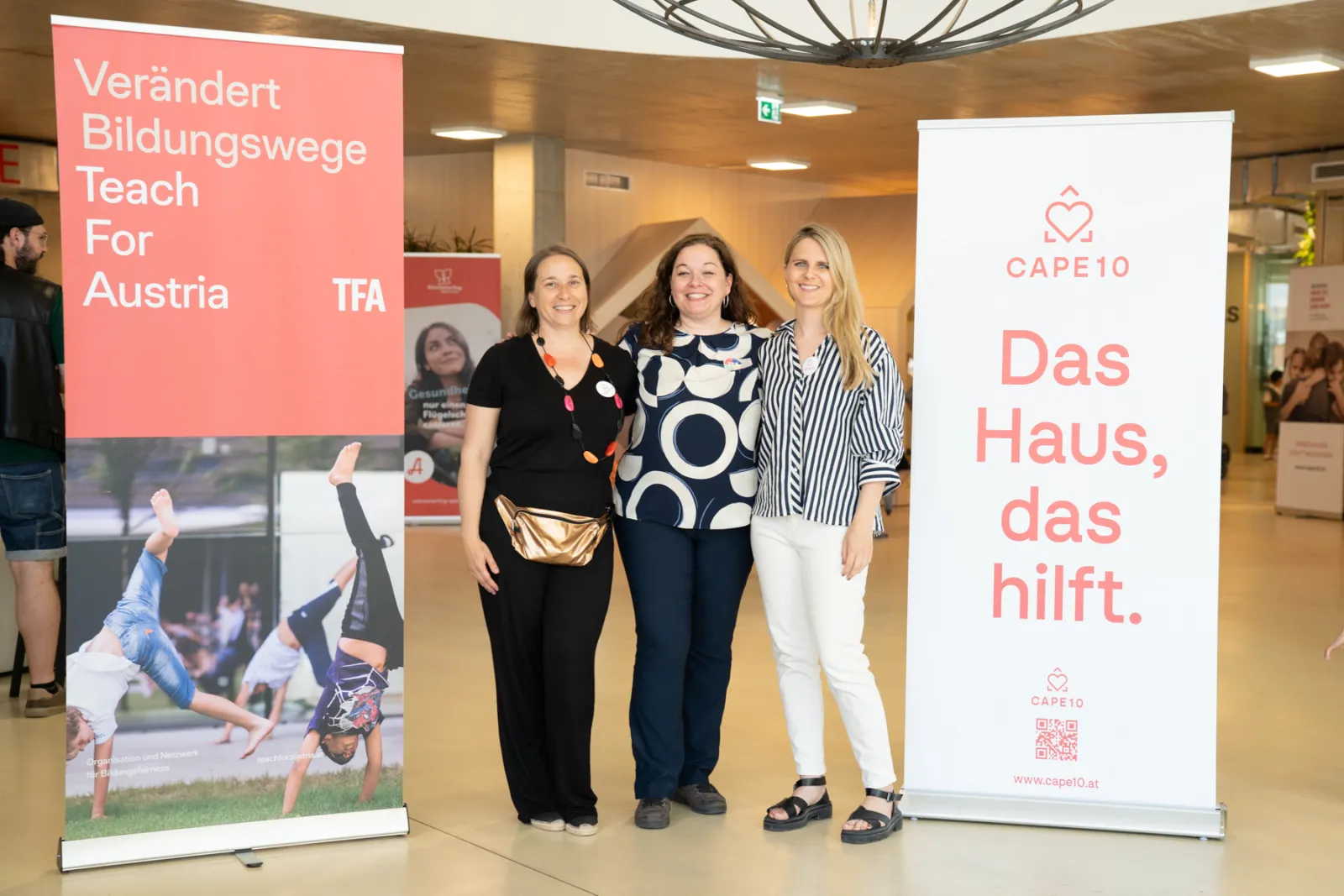 Drei Frauen posieren vor zwei Bannern in einem Innenraum.