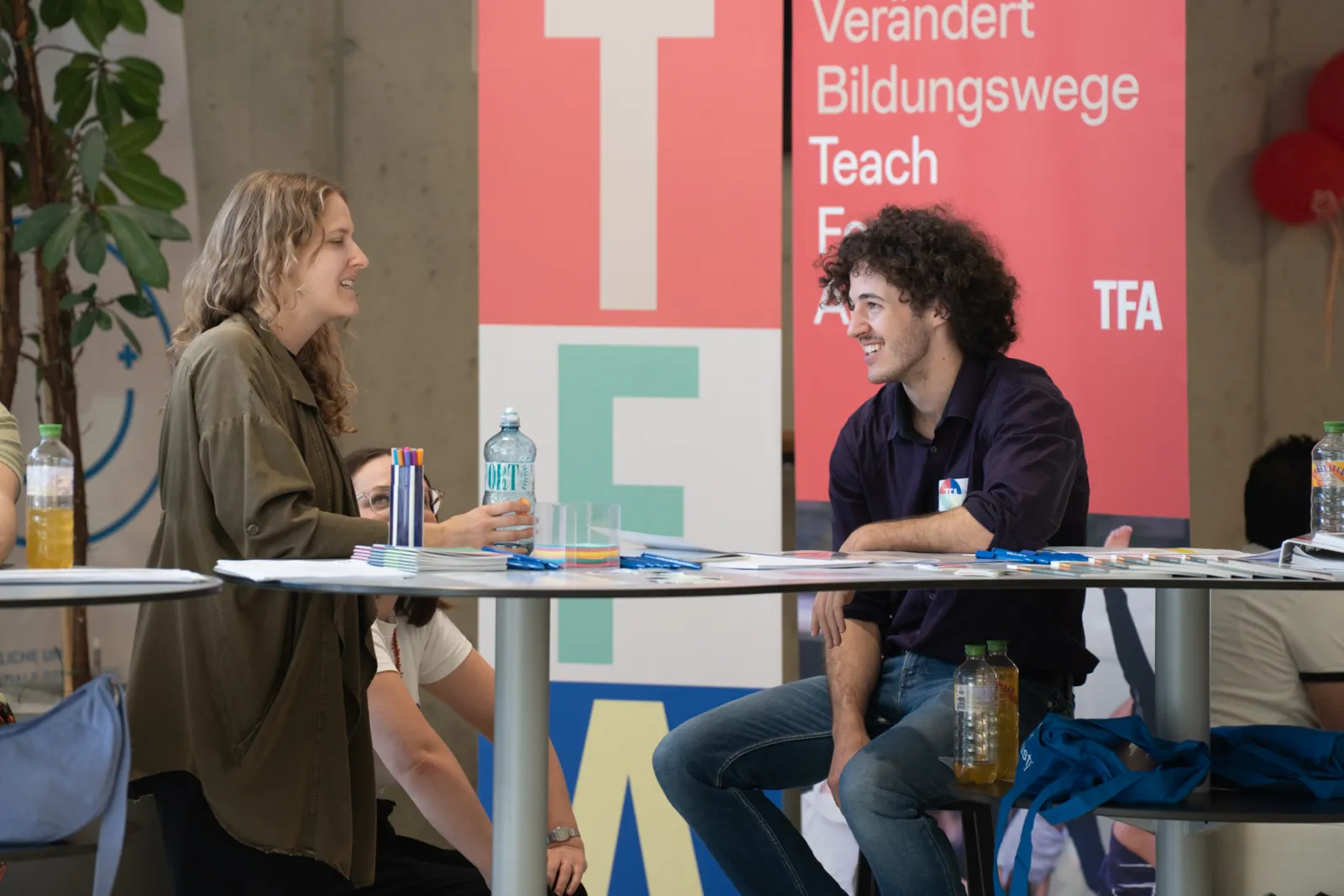 Zwei Personen unterhalten sich an einem Tisch mit Unterlagen, Stiften und Wasserflaschen; im Hintergrund Banner einer Veranstaltung.