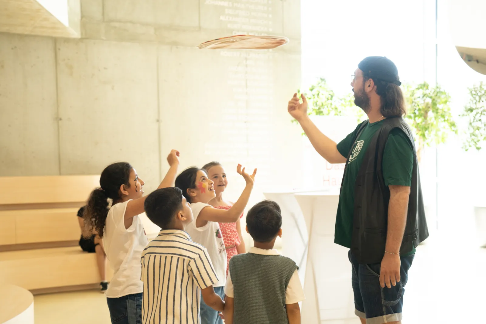 Eine erwachsene Person wirft einen Frisbee zu einer Gruppe von Kindern in einem hellen Innenraum mit Betonwänden und Pflanzen im Hintergrund.