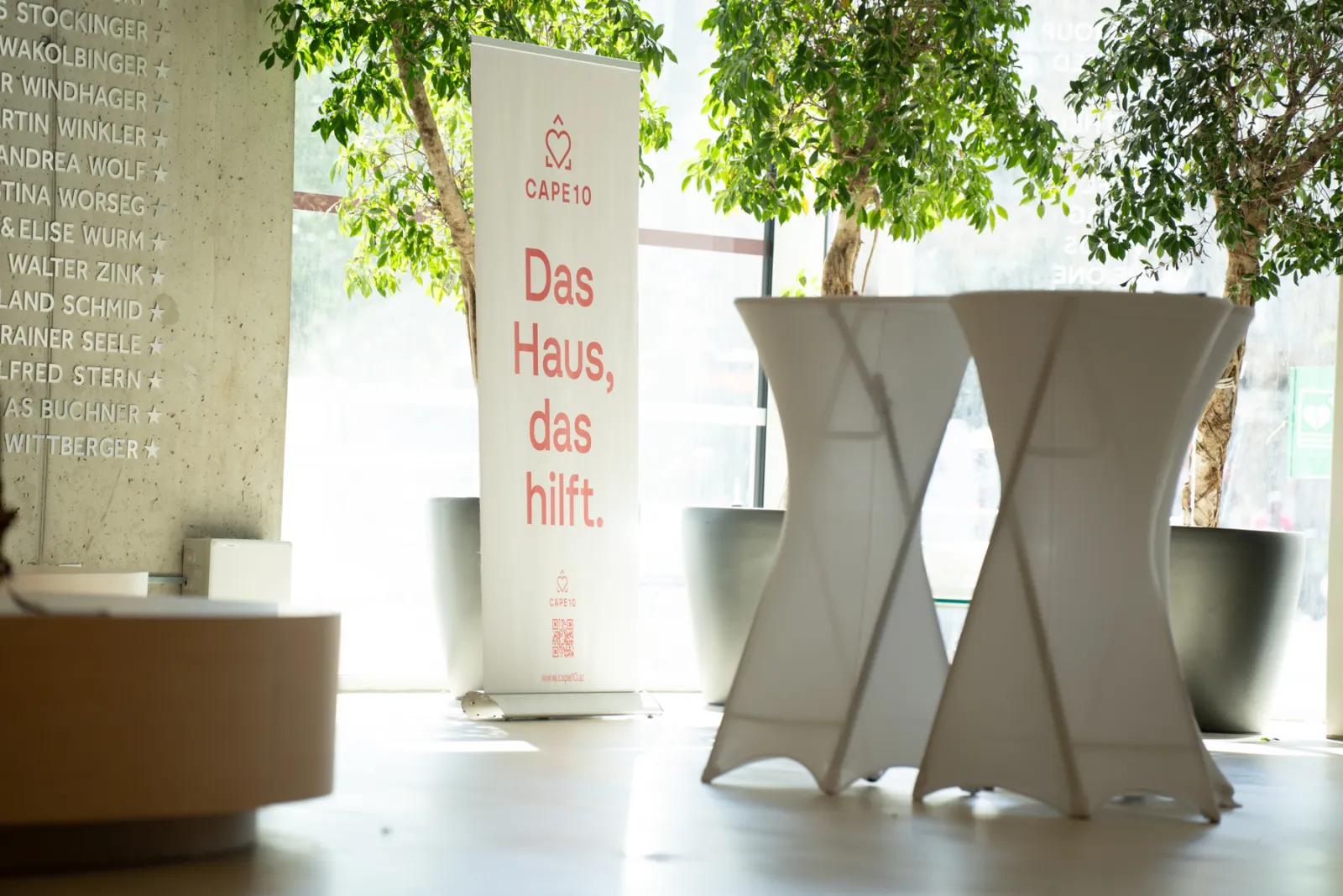 Vertikales Werbebanner mit Text 'Das Haus, das hilft' im hellen Innenraum, flankiert von Pflanzen und modernen Stehtischen.