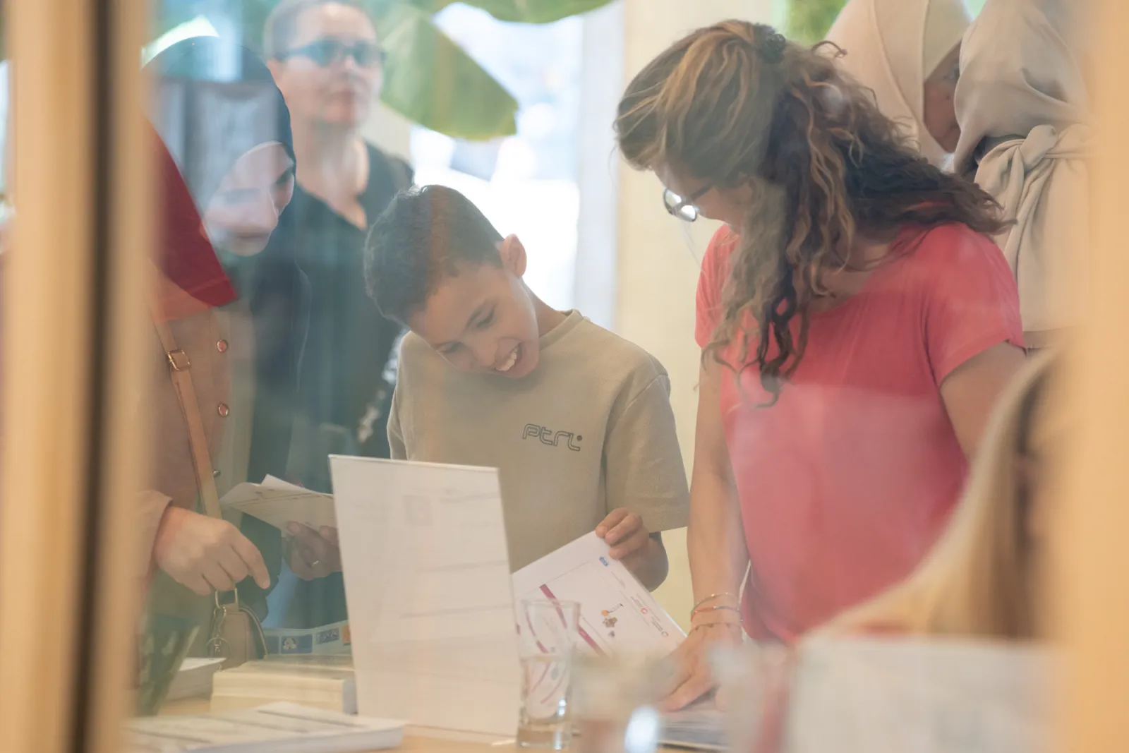 Junge Person blättert lächelnd durch Unterlagen, von Erwachsenen am Infostand umgeben.