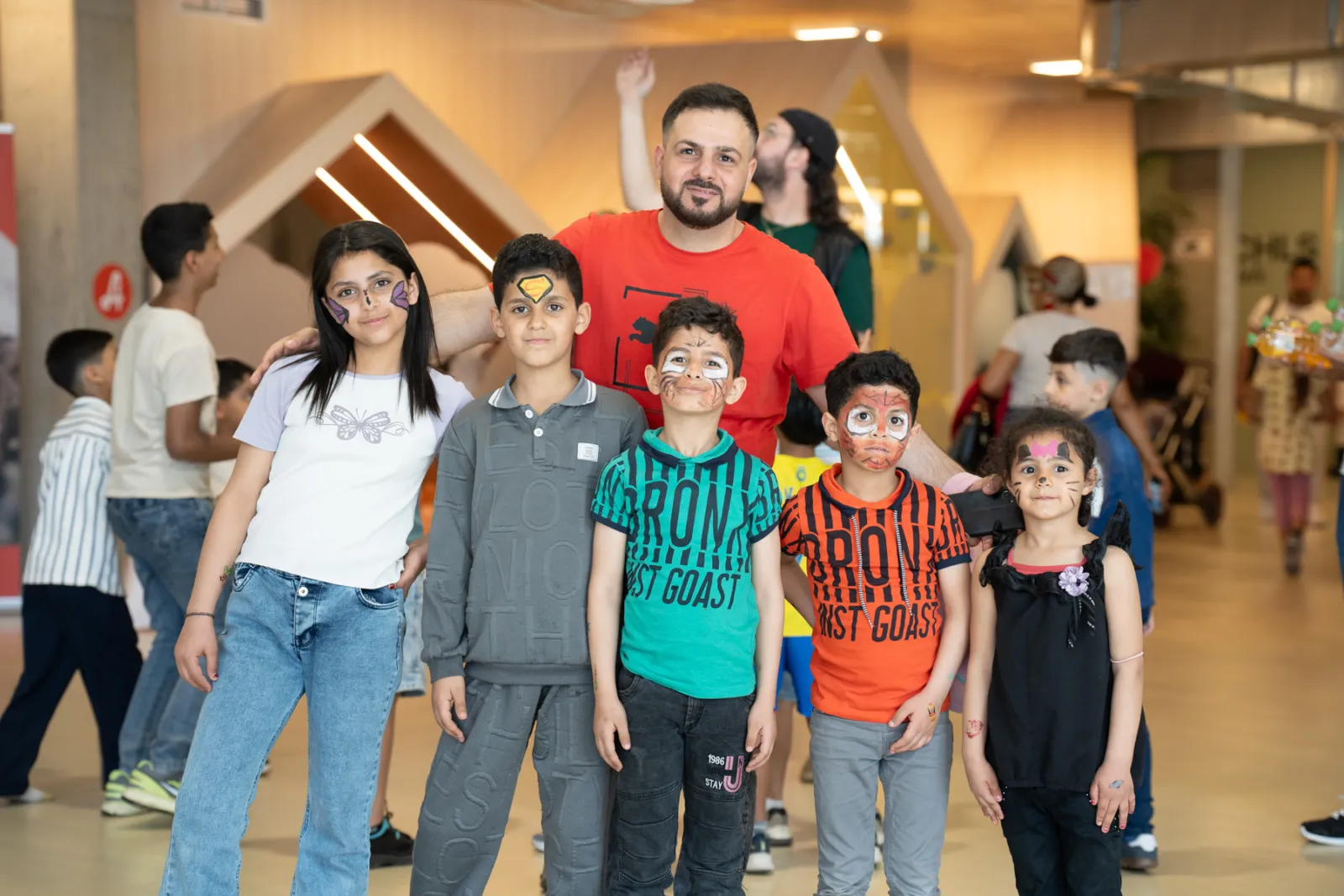 Gruppe von fünf Kindern mit Gesichtsbemalung posiert für Foto; erwachsener Mann im roten T-Shirt steht dahinter; Indoor-Veranstaltung.