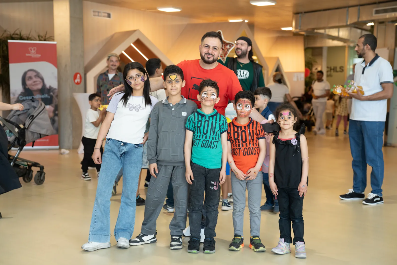 Fünf Kinder mit Gesichtsbemalung posieren für ein Foto; dahinter eine erwachsene Person in rotem Oberteil; Indoor-Veranstaltung in Halle.