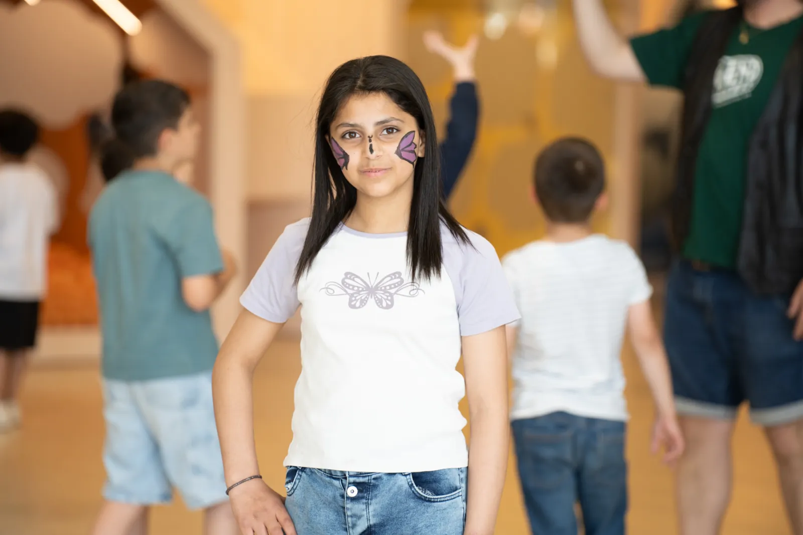 Kind mit Schmetterlings-Make-up posiert im Flur, Hintergrund mit weiteren Kindern.
