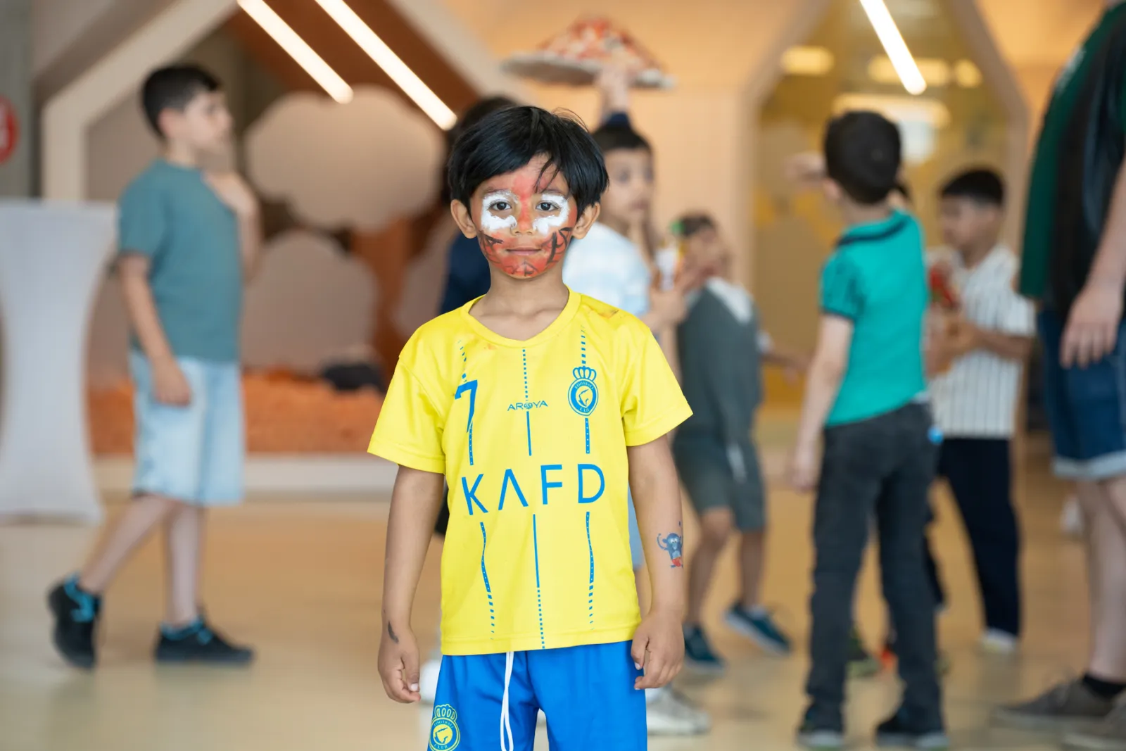 Kind mit Gesichtsbemalung im gelben Trikot steht in einem Indoor-Spielbereich, umgeben von weiteren Kindern.