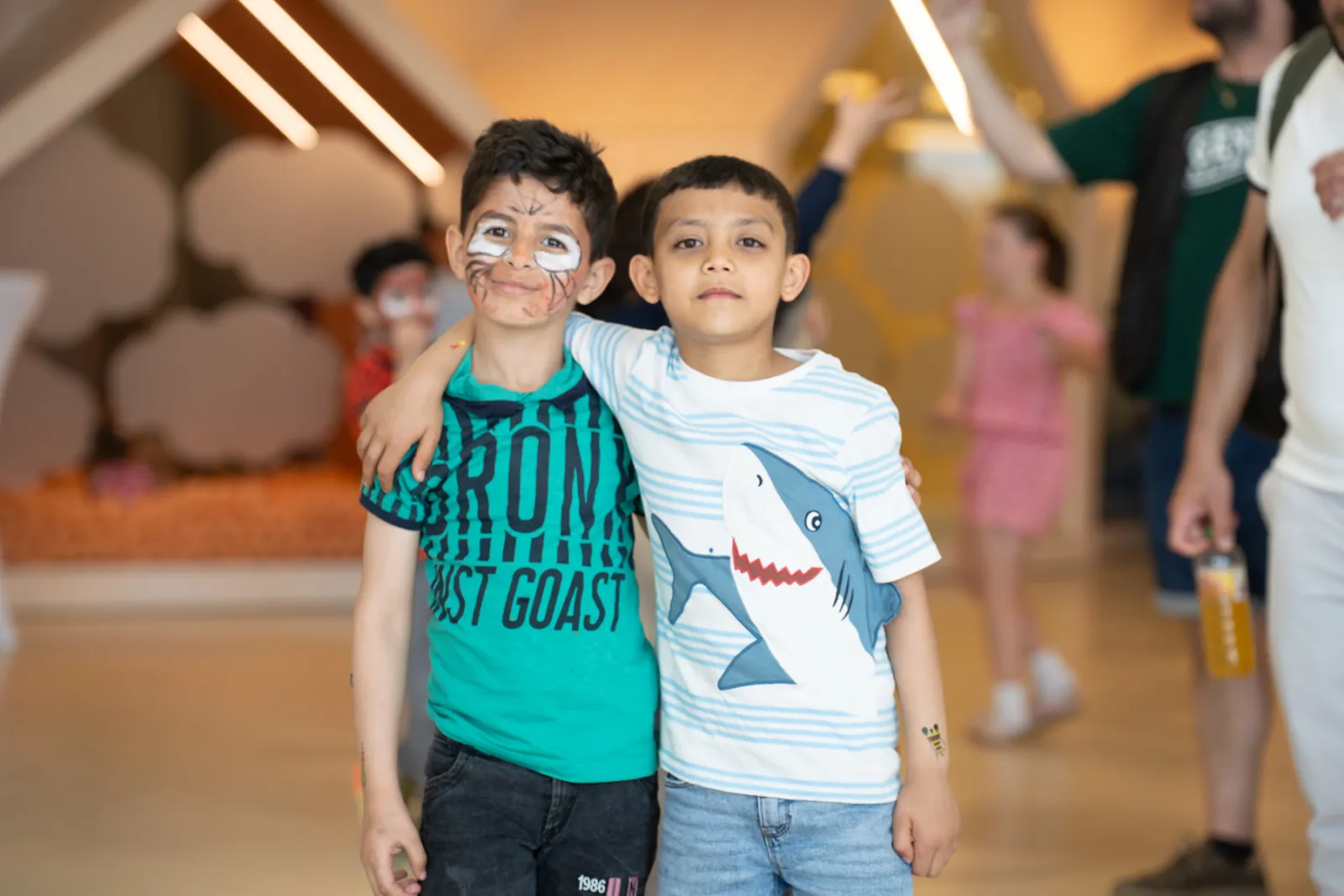 Zwei Jungen stehen mit Arm um die Schultern in einem hellen Innenbereich einer Veranstaltung; der linke hat Gesichtsbemalung.