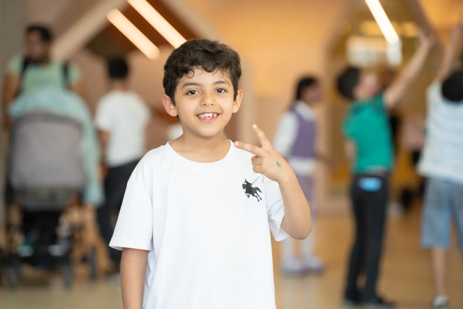 Kind lächelt und zeigt Peace-Zeichen in belebtem Innenraum, Hintergrund mit weiteren Kindern.