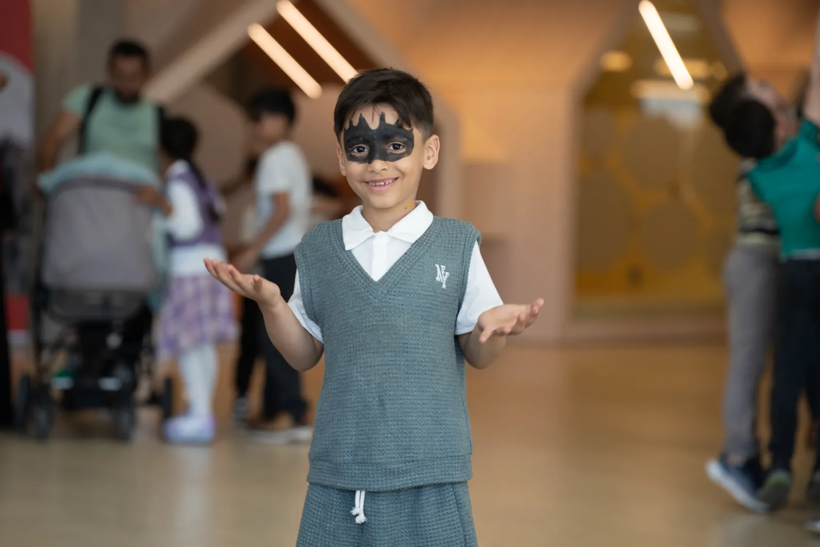 Kind mit Batman-Maske, graues Weste über weißem Polohemd, hebt die Hände, steht in einer Einkaufspassage
