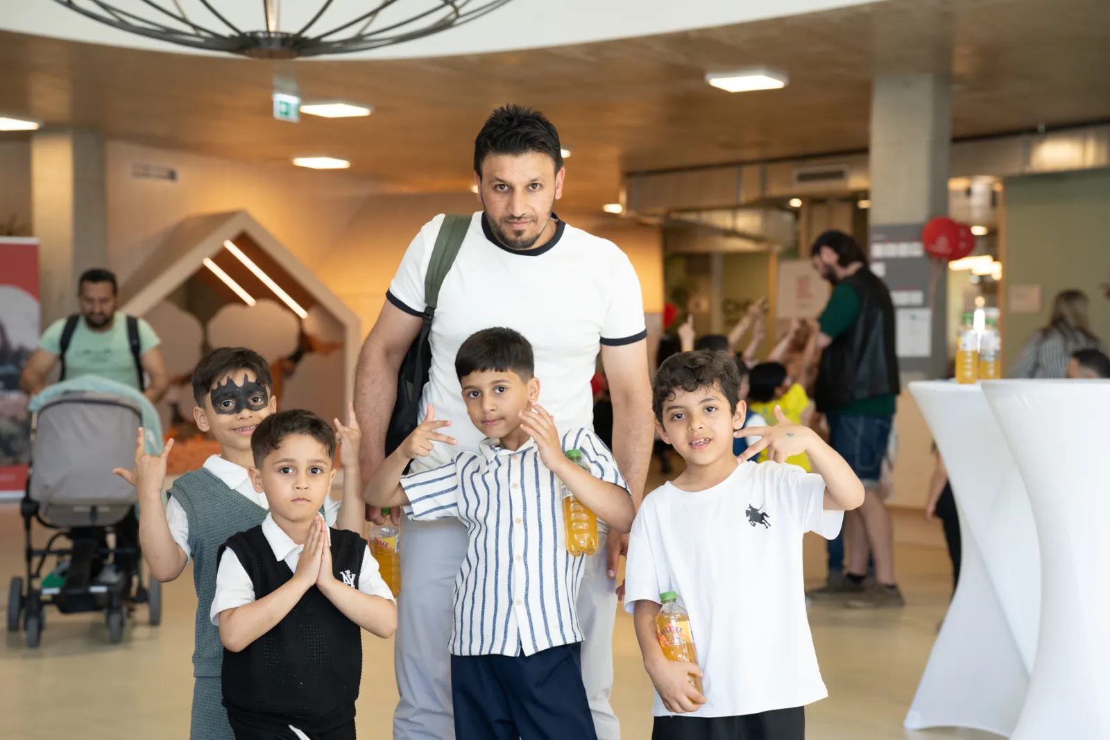 Erwachsener Mann mit vier Jungen posiert in hellem Innenbereich; Kinder halten Getränkeflaschen, einer zeigt Handzeichen, einer trägt Maske