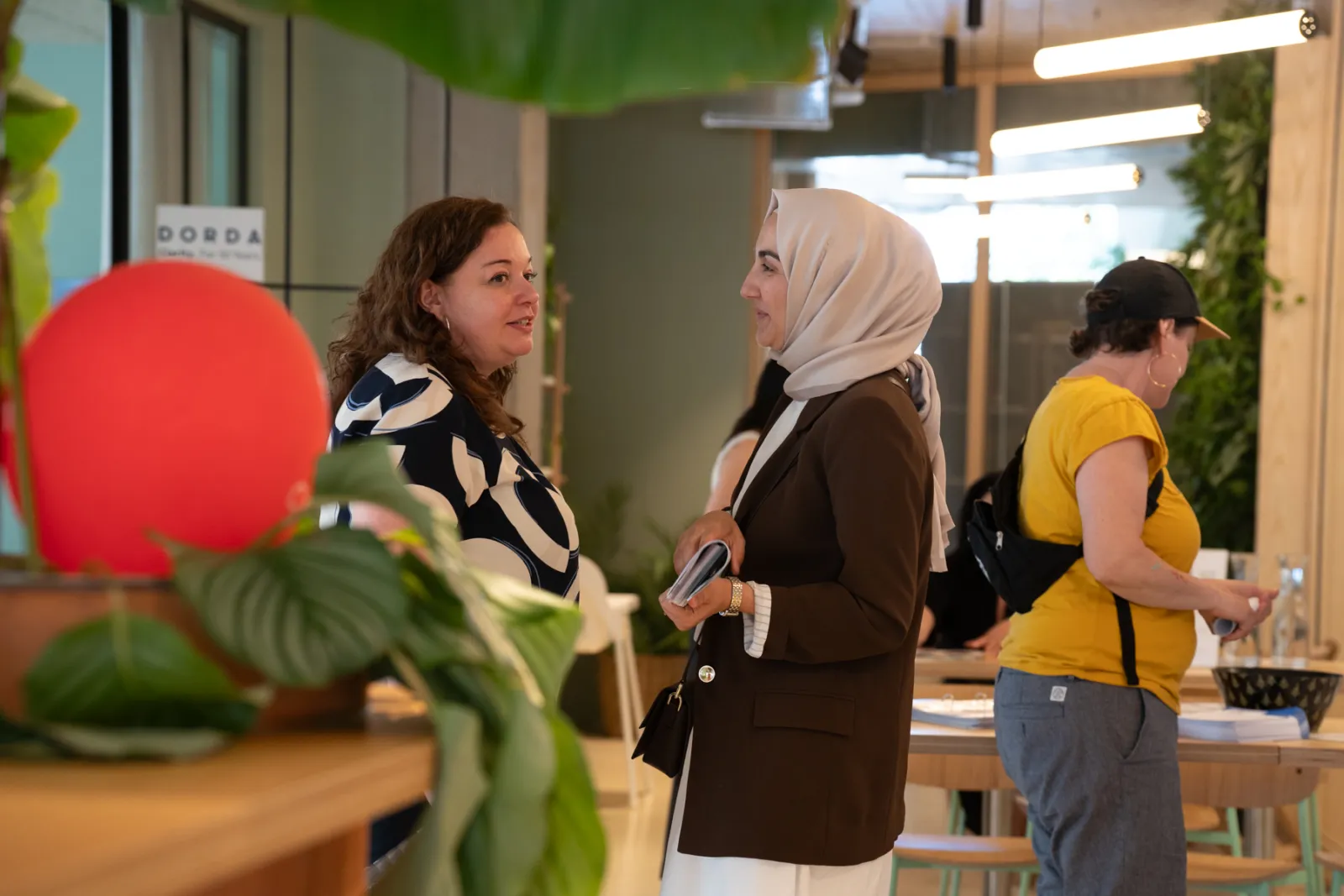 Zwei Personen im Gespräch in einer modernen Café-Lounge; eine Person mit Hijab in braunem Blazer, die andere in gemustertem Oberteil.