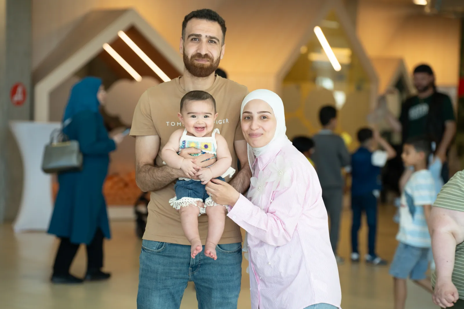 Vater hält Baby; Mutter im Kopftuch daneben, vor modernem Innenraum mit weiteren Besuchern.