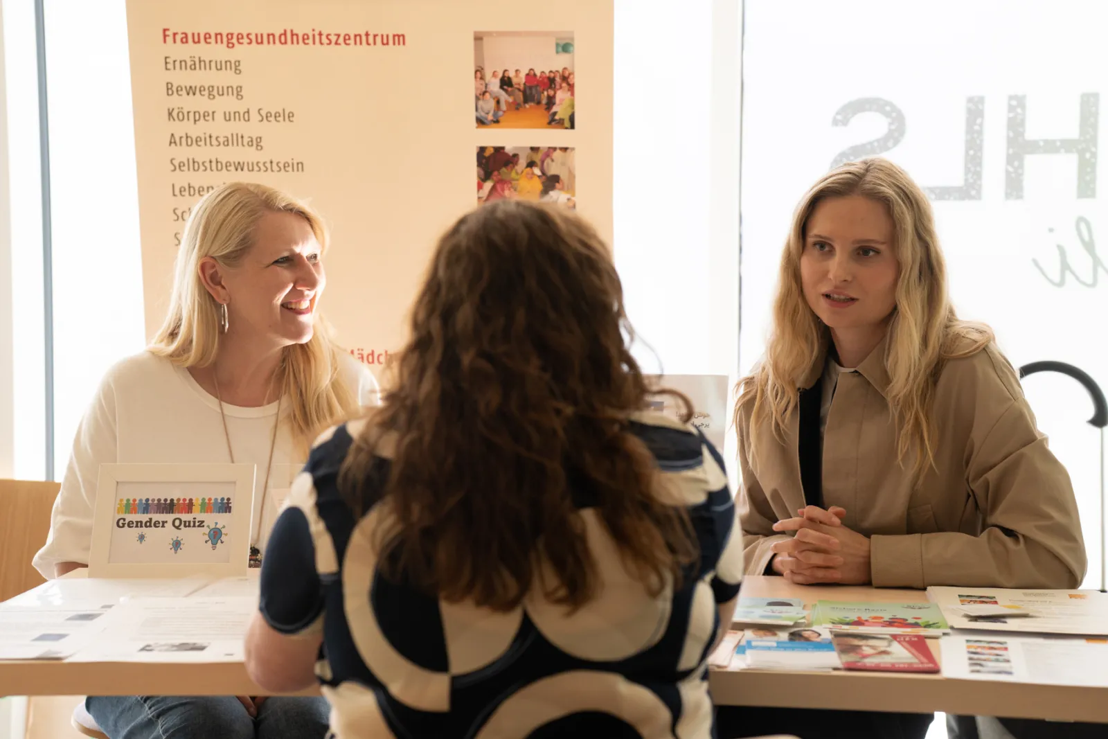 Zwei Frauen beraten eine Person an einem Informationsstand; Tisch mit Schild 'Gender Quiz' im Gesundheitszentrum.