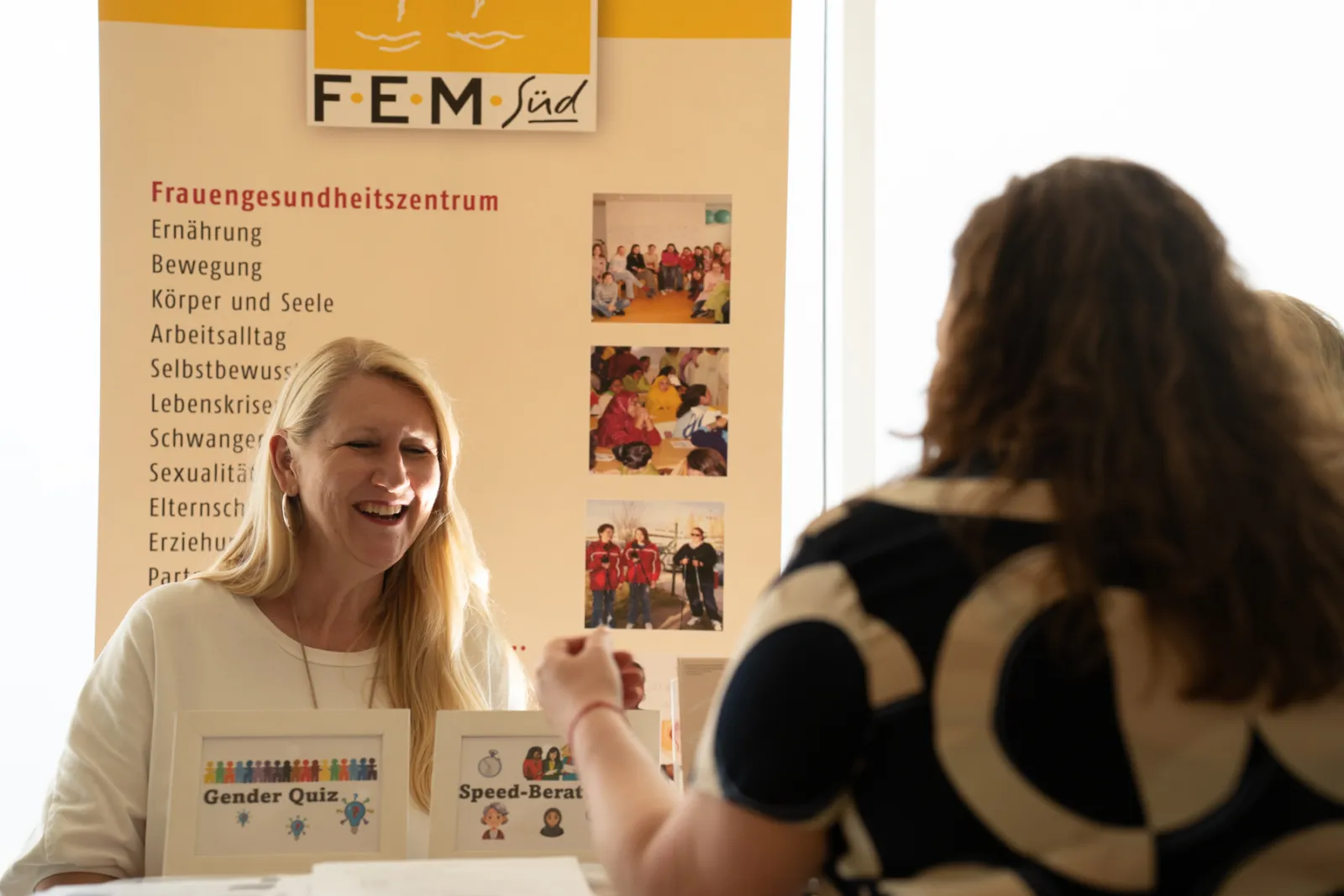 Lächelnde blonde Frau hinter Infostand des Frauen-Gesundheitszentrums; Banner mit Themenliste im Hintergrund.